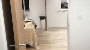 Un dormitorio con cama, mesita de noche y tocador. Se ve una puerta que da a un pasillo, donde se distingue una zona de cocina al fondo.