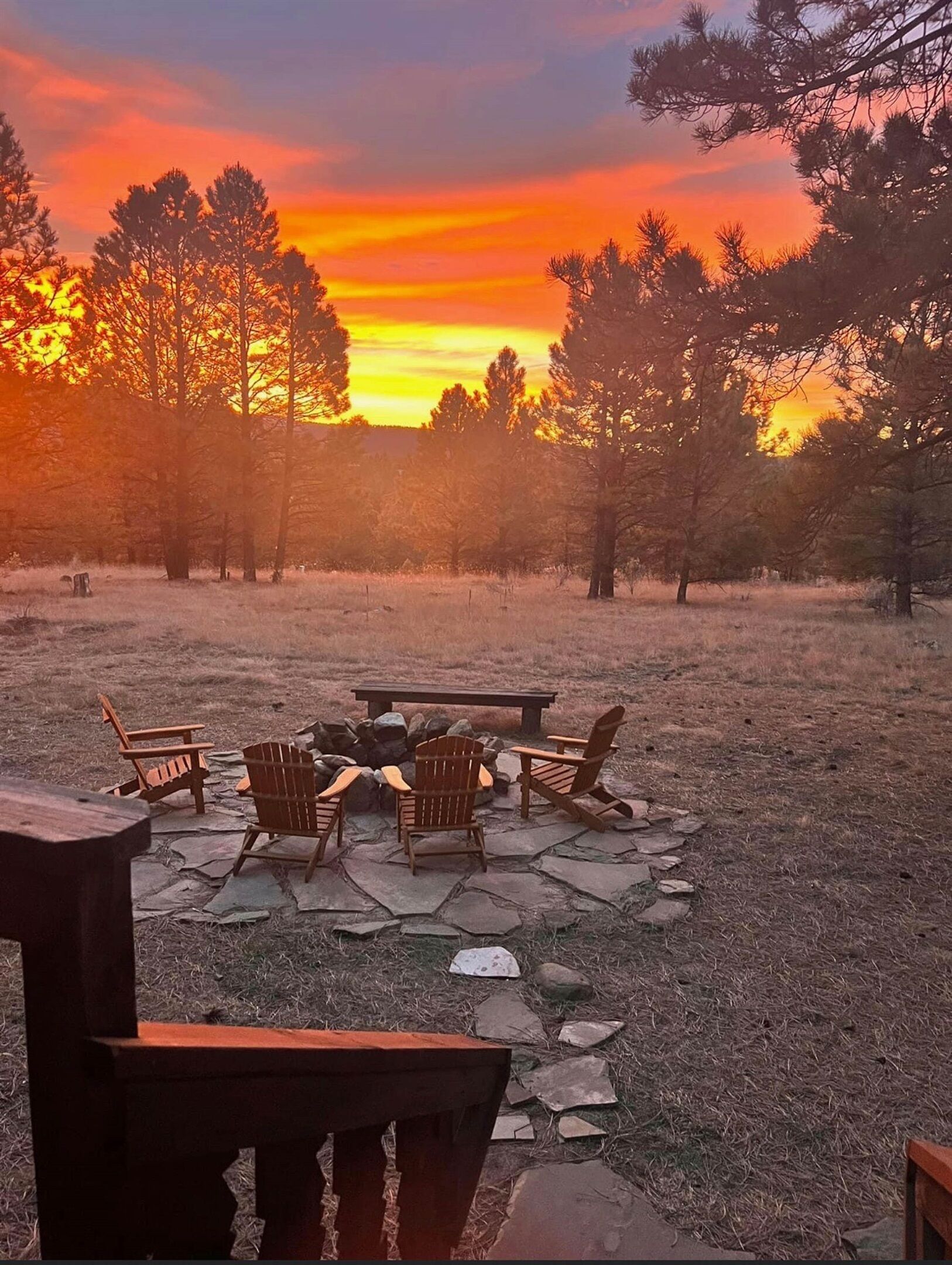 Een terras met een vuurkorf van natuursteen en Adirondack-stoelen, omringd door een grasveld en bomen, tijdens de zonsondergang.