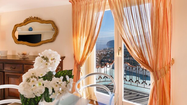 Una stanza con vista sulla città attraverso una porta finestra, uno specchio sulla parete e un vaso con fiori bianchi su un tavolo.