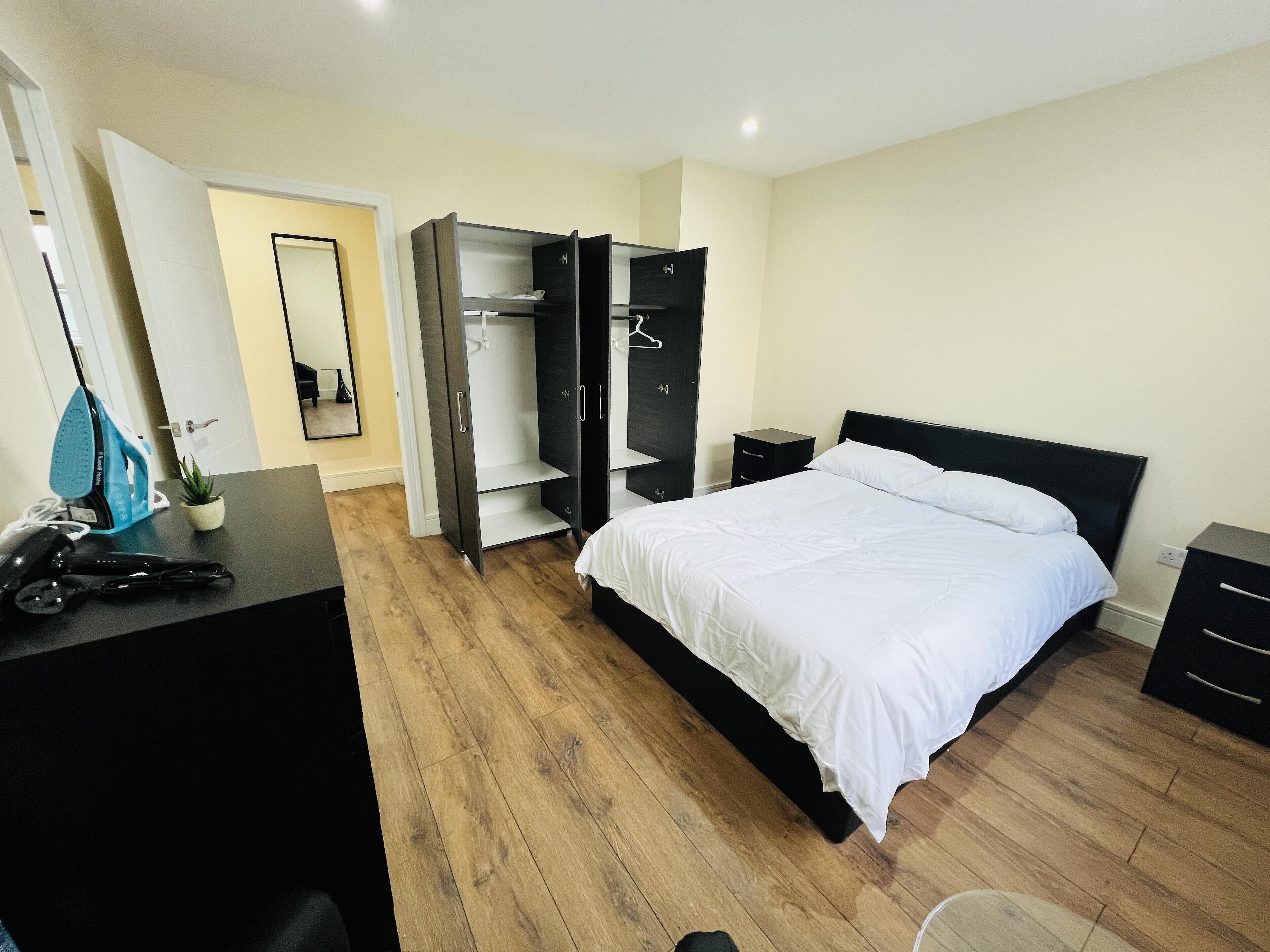 A bedroom with a bed, a wardrobe, a desk with an iron, and a small plant.