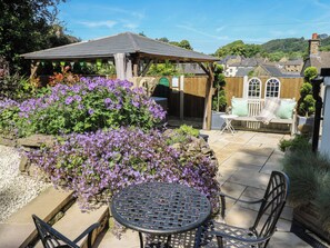 A patio with a round metal table and chairs, surrounded by flowering plants and a wooden pergola.