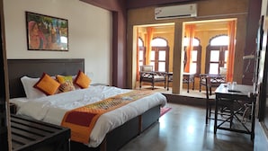 A hotel room with a large bed, a desk, and a chair. There is a painting on the wall and an air conditioning unit above the door. - Hotel Sohandeep (Jaisalmer)