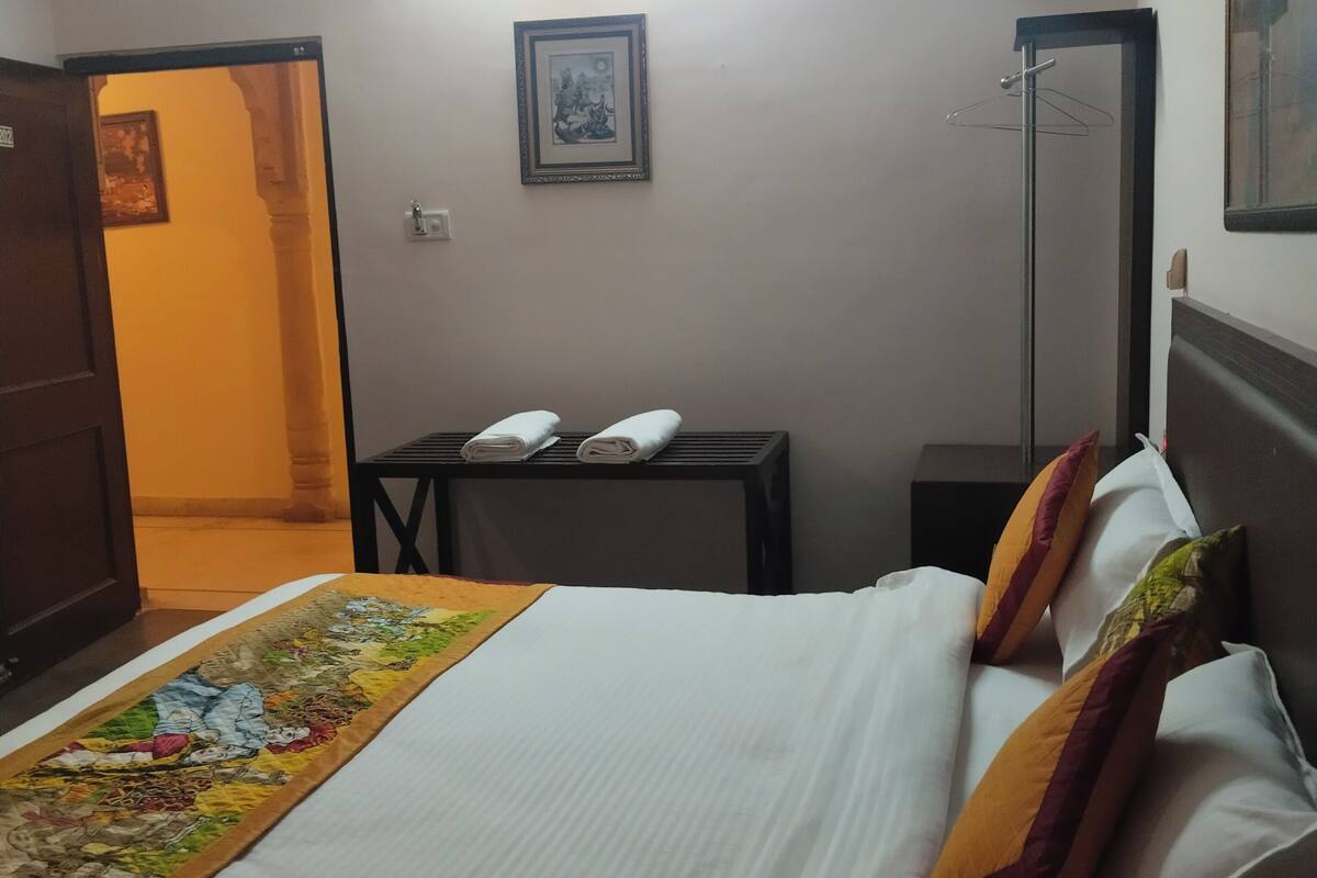 A hotel room with a neatly made bed, a small table with a lamp, a hanging coat rack, and a framed picture on the wall.