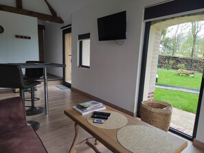 Ein modernes Wohnzimmer mit einem Holztisch, einem an der Wand montierten Fernseher und Blick auf einen Garten durch eine Glastür.
