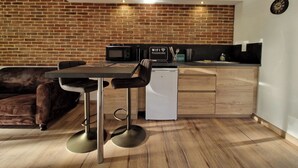 A compact kitchen with a brick wall, a microwave, a coffee machine, and a small refrigerator. There is a wooden dining table with two chairs and a sofa in the living area.