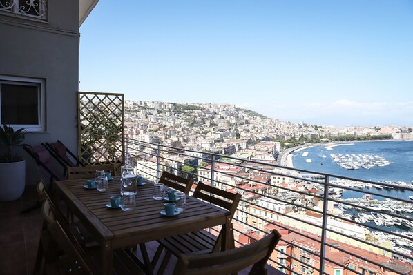A balcony with a wooden table set for dining, overlooking a coastal cityscape with buildings and a marina.
