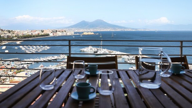 Une table avec des tasses vides et un pichet en verre, donnant sur une marina où sont amarrés des bateaux, avec une montagne qui se profile à l’horizon.