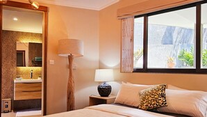 Ein Hotelzimmer mit Bett, Nachttisch mit Lampe, Fenster mit Vorhängen sowie Bad mit Spiegel und Waschbecken.