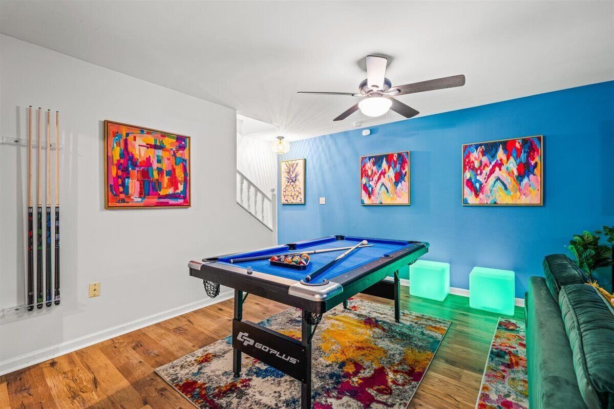 A room with a pool table, colorful abstract paintings, a ceiling fan, and a green couch.