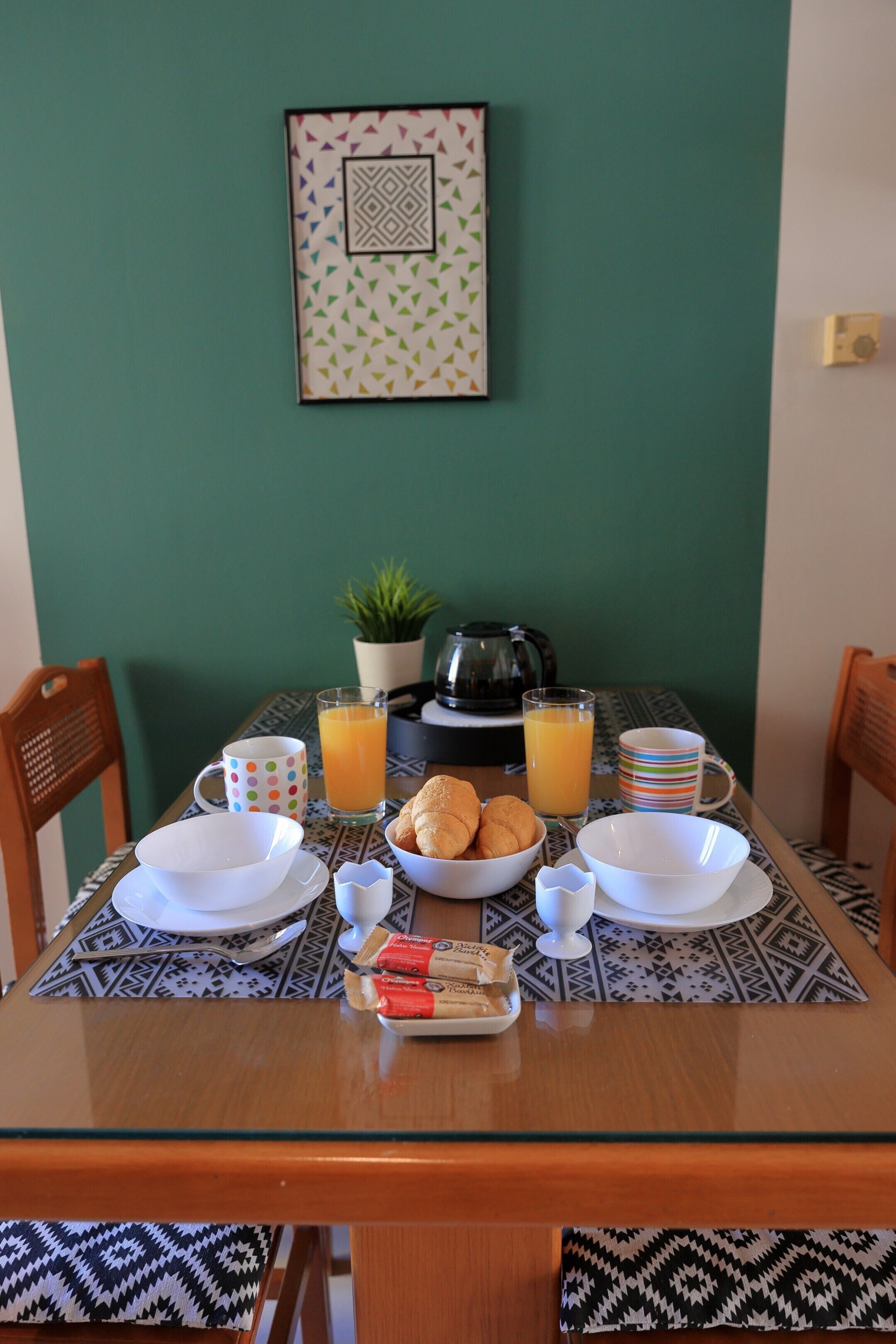A breakfast table with plates, cups, a bowl of croissants, a small tray of pastries, and a coffee maker.