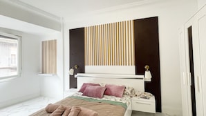 A modern bedroom with a large bed, a bedside table, a nightstand, a built-in headboard with vertical slats, and a large window. - Apartament Irun (Irun)