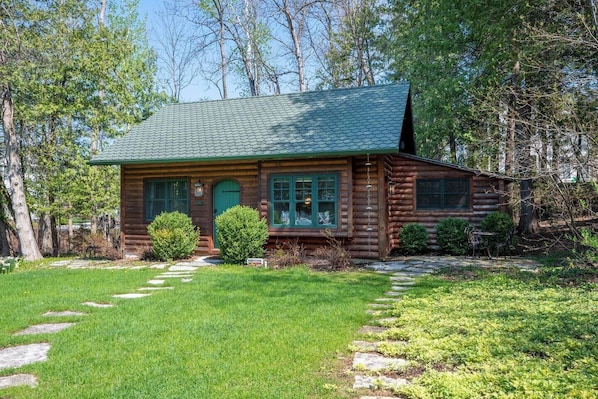 A log cabin with a green roof, surrounded by a stone pathway and lush greenery.
