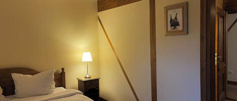 A bedroom with a bed, a nightstand with a lamp, a wooden ceiling beam, a framed picture on the wall, and a door leading to another room.