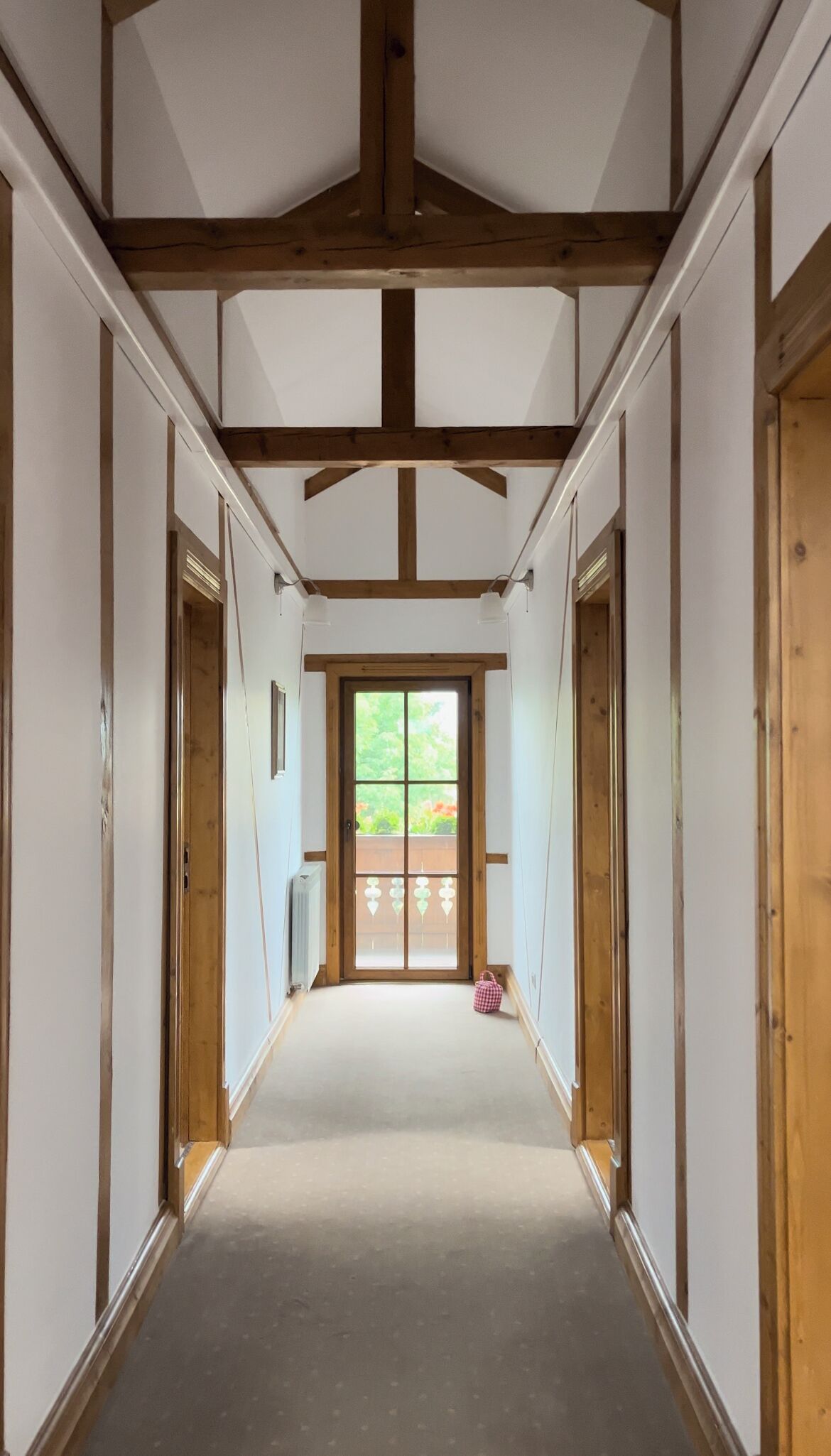 Un couloir étroit avec des poutres en bois au plafond, des murs blancs et une grande porte en bois au fond.
