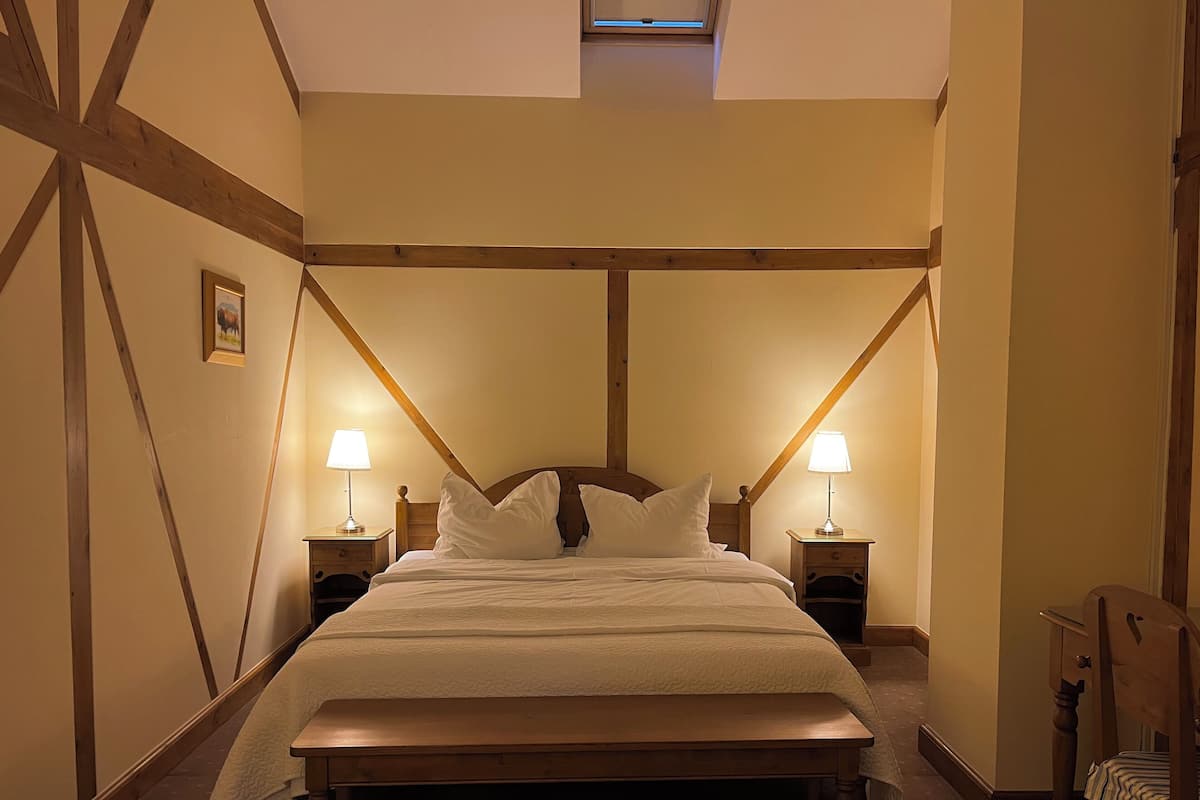 Un dormitorio con una cama grande, dos mesitas de noche con lámparas, un banco de madera y vigas de madera a la vista.
