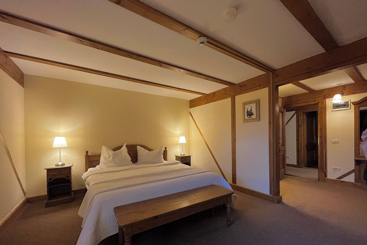 Un dormitorio con una cama de madera, dos mesitas de noche con lámparas, un banco y una puerta que da a otra habitación.