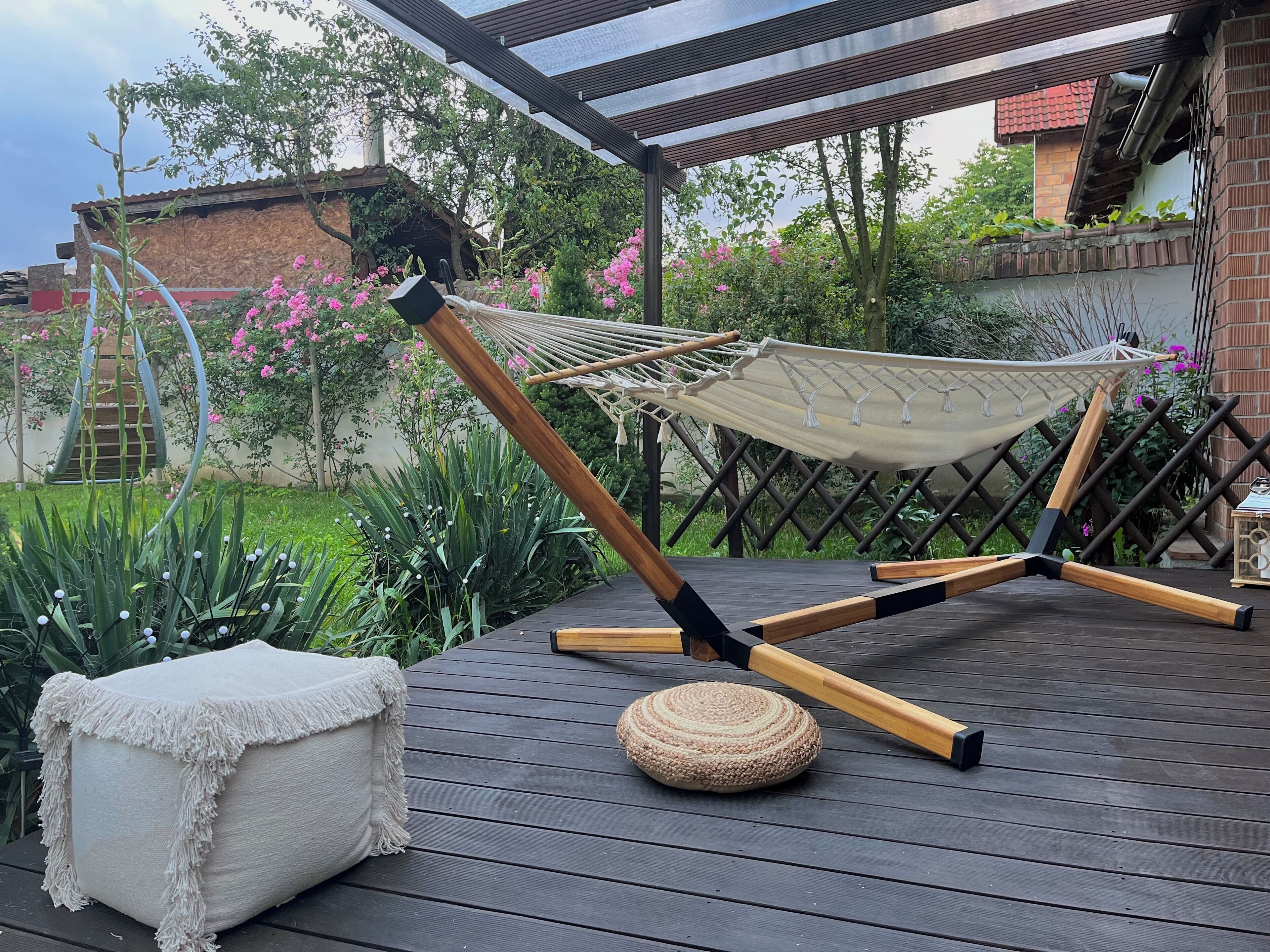 Uma rede sobre um deck de madeira, com uma almofada branca e um banquinho de fibra natural por perto.