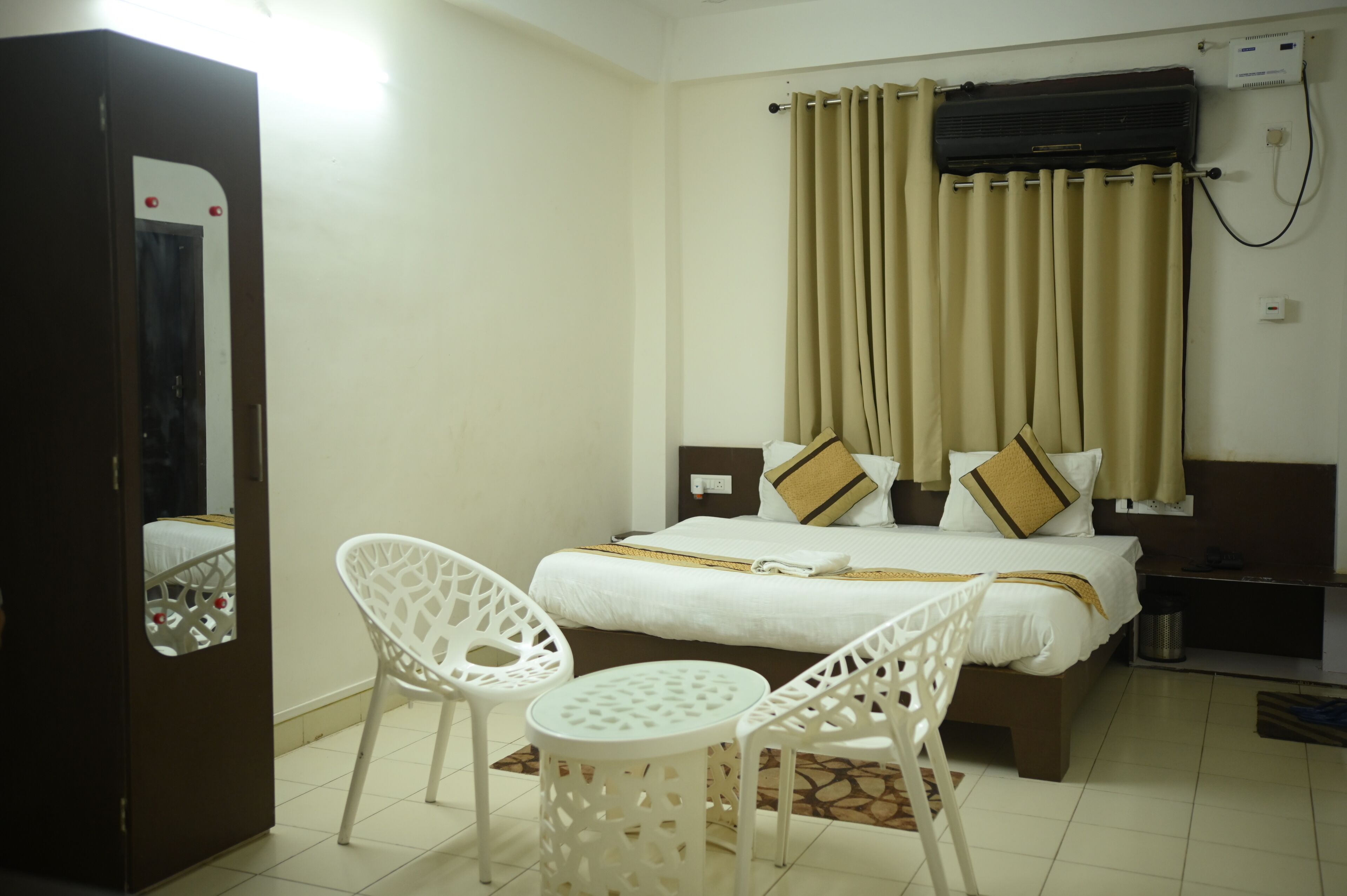 A hotel room with a bed, two white chairs, a small round table, a curtain, and a wall-mounted air conditioner.