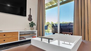 A modern living room with a large TV, a guitar, and a balcony with outdoor furniture. - Walk to Gruene-Guadalupe Riverfront-River Access-Stylish Texas Music Retreat (New Braunfels)