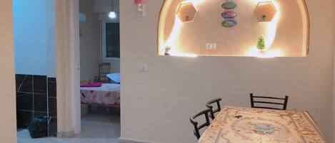 Un comedor con una mesa y sillas, una cocina visible al fondo, y una pared decorativa con un aplique con forma de casa.