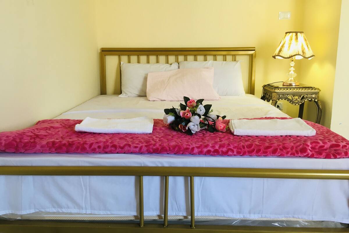 A neatly made bed with a pink floral bedspread and white linens, a bedside lamp, and a bouquet of flowers on the bed.