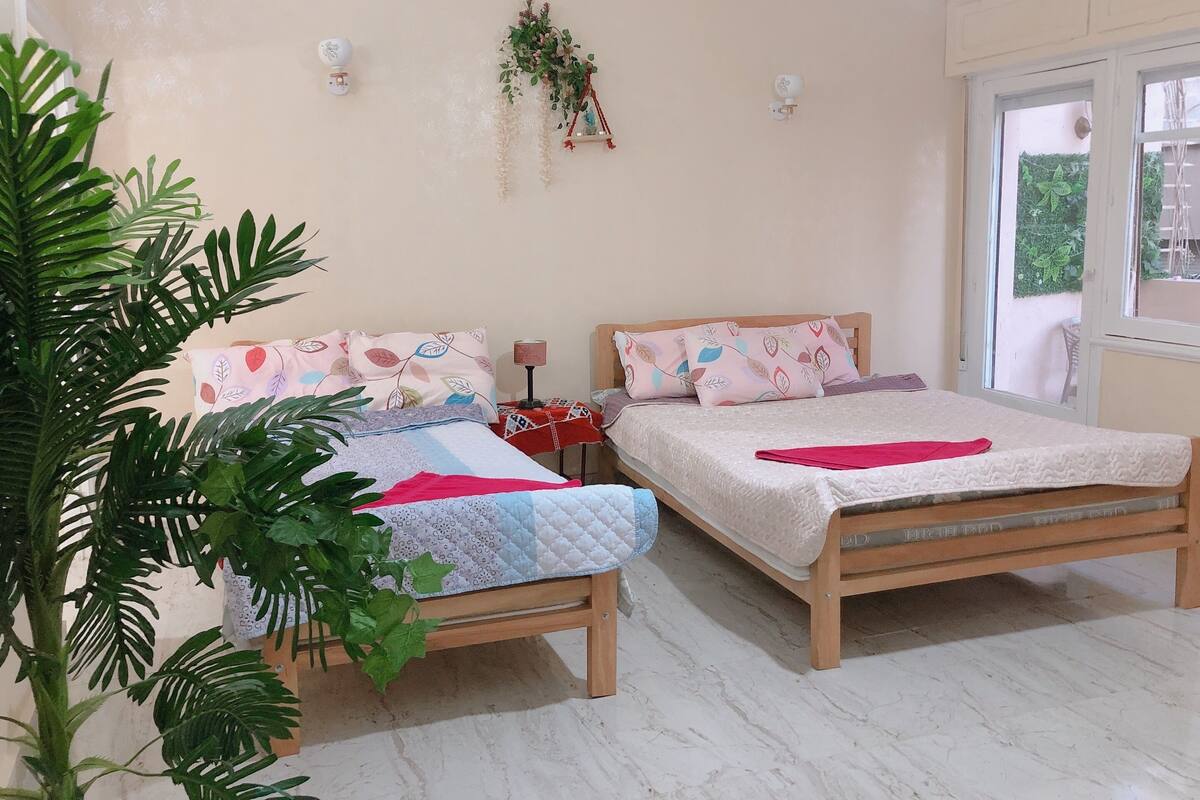 A bedroom with two beds, a potted plant, wall-mounted lights, and a window.