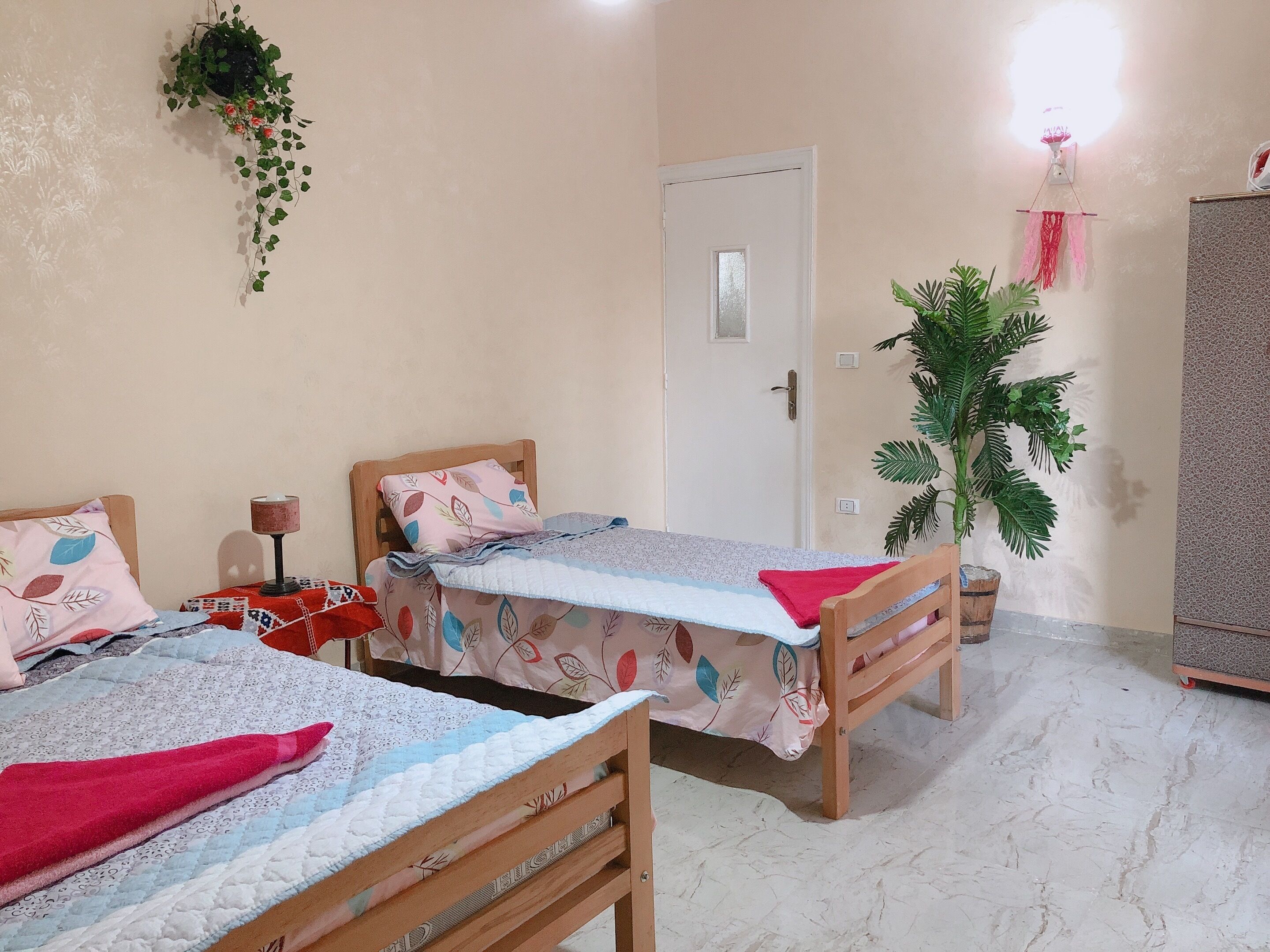 A room with two wooden beds, a small table with a lamp, a potted plant, and a wardrobe.