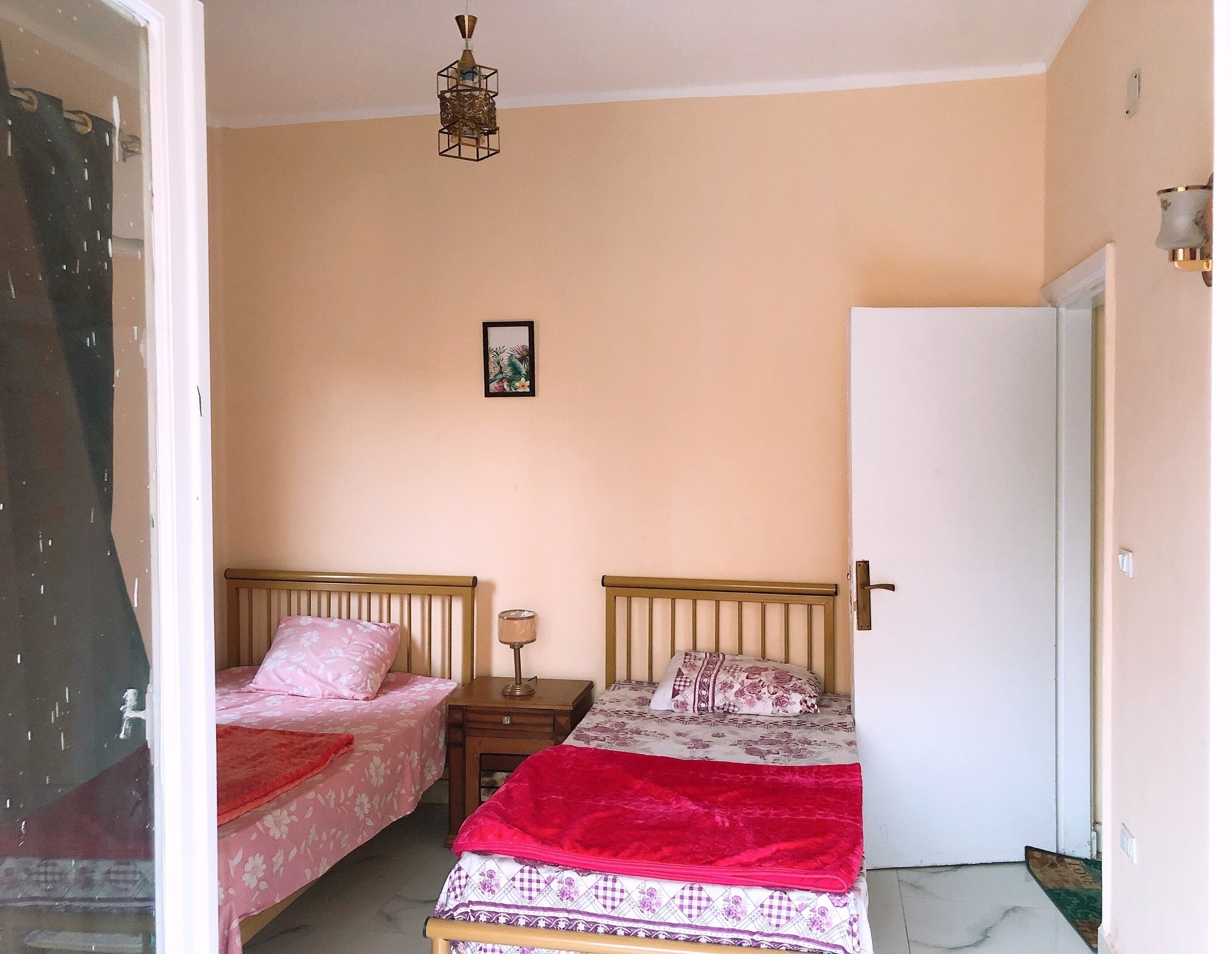 A bedroom with two single beds, a bedside table with a lamp, a framed picture on the wall, and a door to the right.