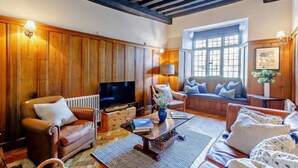 A cozy living room with wooden paneling, a television, comfortable seating, and a large window providing natural light. - Ostlers - beautiful former Coaching Inn with parking and late Sunday checkout. (BURFORD)