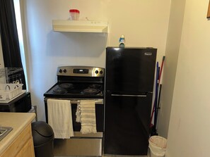 A compact kitchen with a black stove, a refrigerator, and a microwave. There is a small shelf above the stove holding a container and a bottle. - Aria Hospital, I-95, NY ramp (Philadelphia)