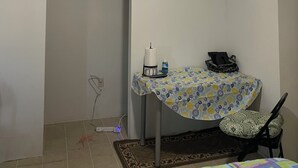 A small room with a ceiling fan, a table with a floral tablecloth, a chair, a framed picture on the wall, and a power outlet on the floor.