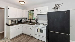 A compact kitchen with white cabinets, a black refrigerator, a microwave, a stove, and a window with blinds.