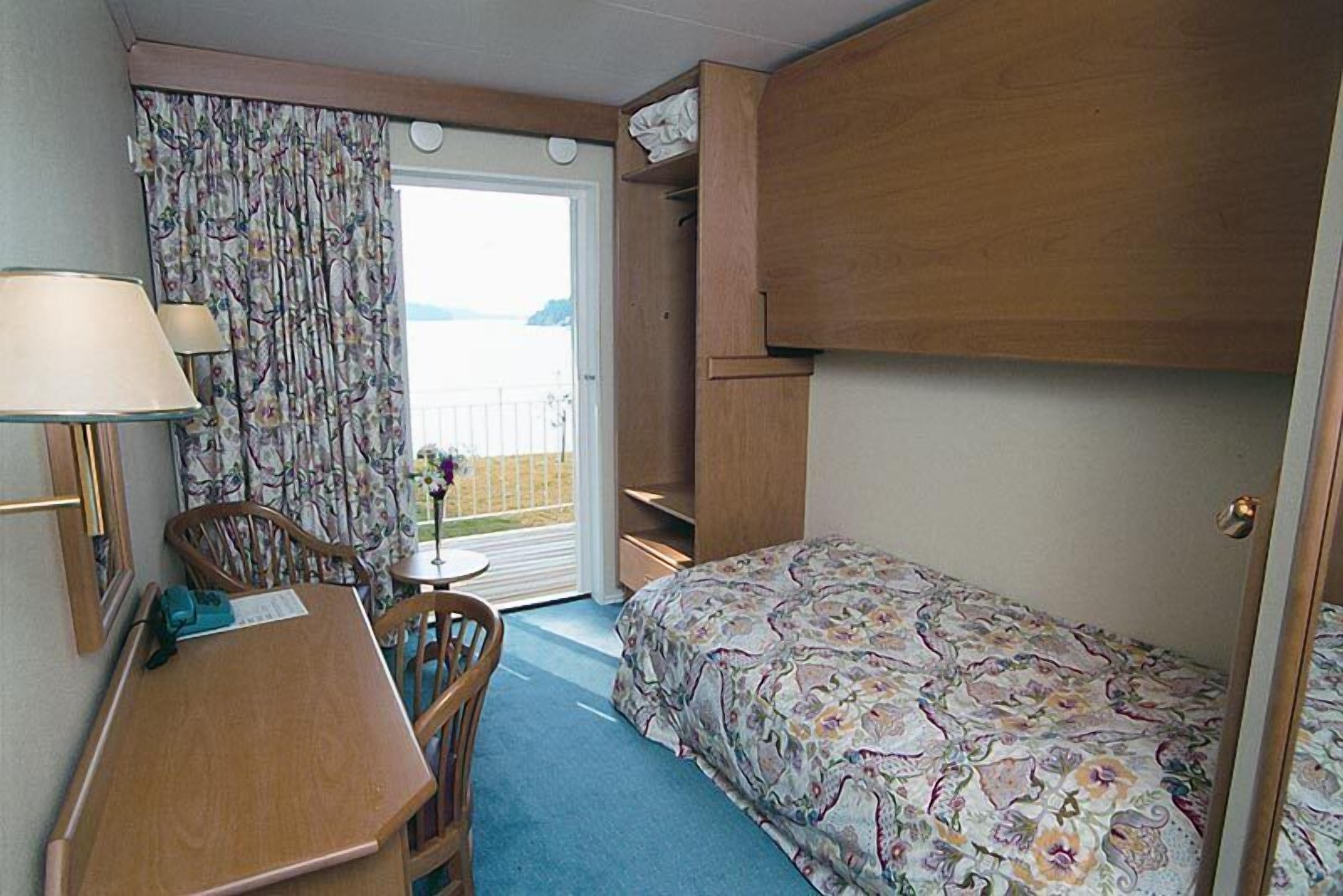 Habitación compacta con cama, escritorio, silla y una pequeña mesa. Tiene una ventana con cortinas, una puerta y vistas al exterior.