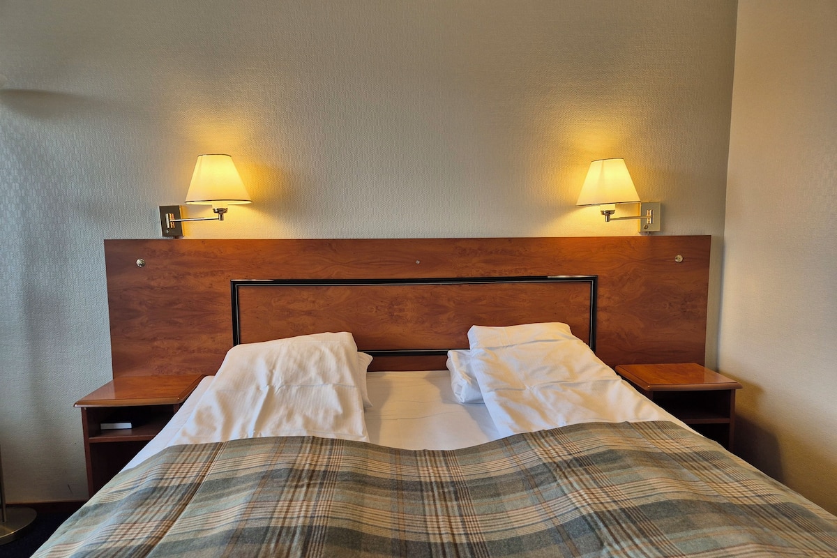 A hotel room with a wooden headboard, two bedside lamps, a plaid bedspread, and a nightstand with a lamp.