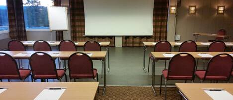 Un salón de conferencias con filas de escritorios y sillas, una pantalla de proyección y una pizarra.