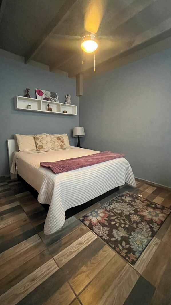 A bedroom with a bed, a nightstand, a lamp, a shelf with decorative items, and a ceiling light.