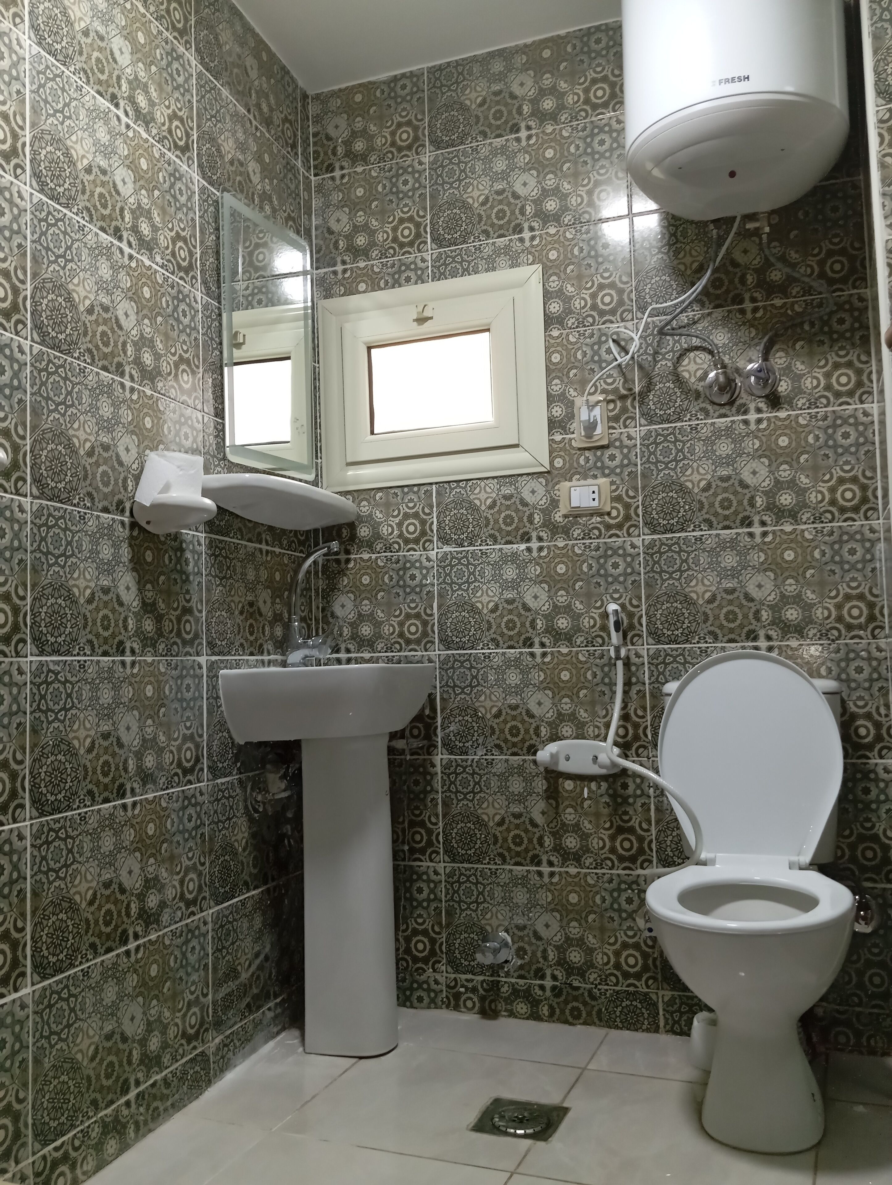 Casa de banho com azulejos estampados, lavatório assentado, sanita, espelho e termoacumulador no teto.