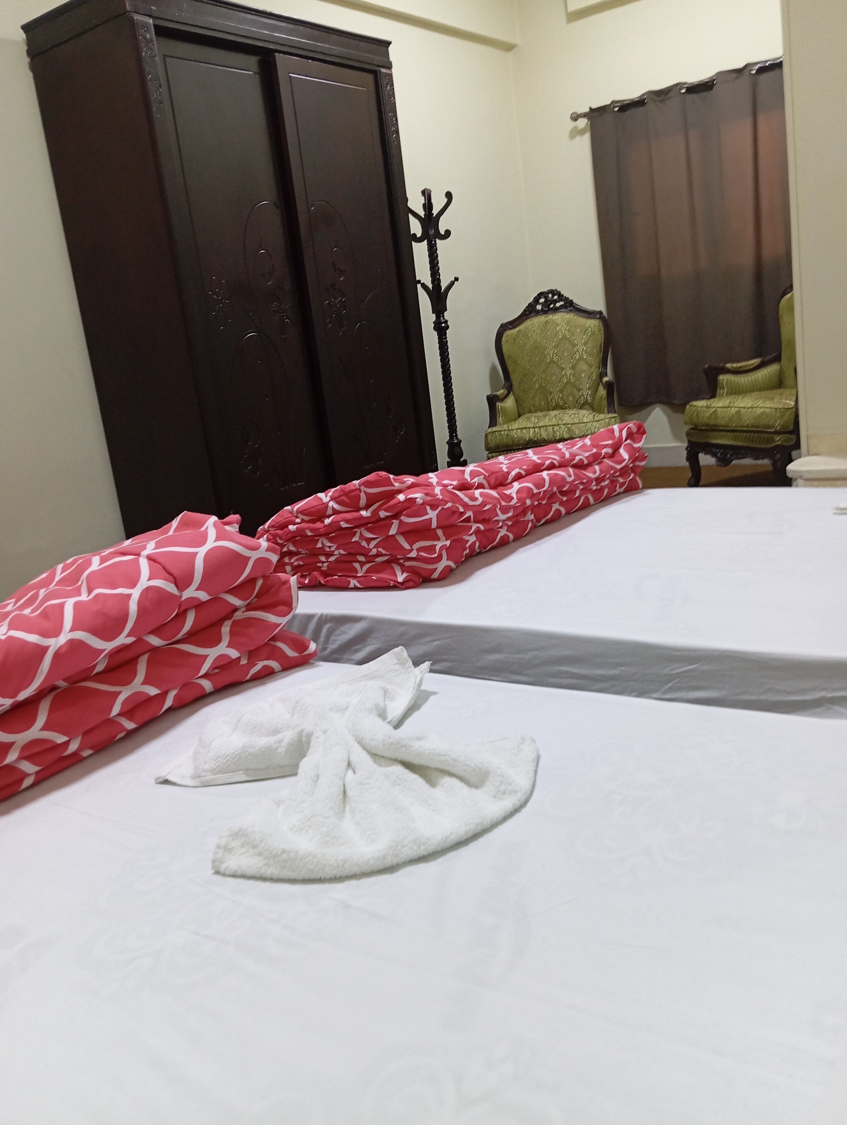 A neatly made bed with a white and red patterned blanket, a white towel, and a dark wooden wardrobe in a room with a green chair and a dark curtain.