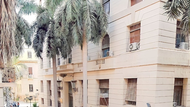 Ein Balkon mit Korbstuhl und kleinem Tisch, mit Blick auf eine Straße mit Gebäuden und Palmen.