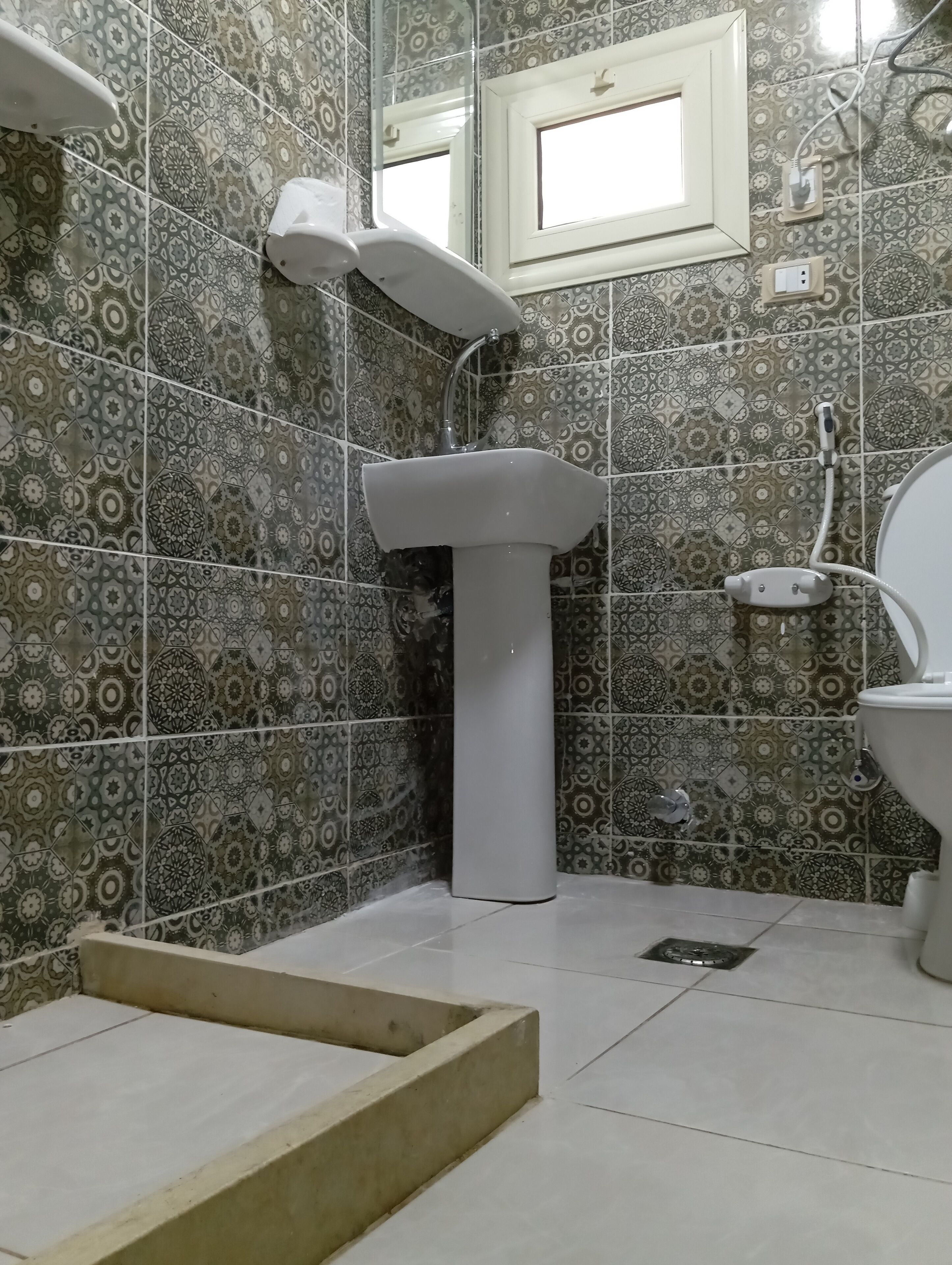 Ein Badezimmer mit gemusterten Fliesen, einem Standwaschbecken, einer Toilette und einem Fenster.