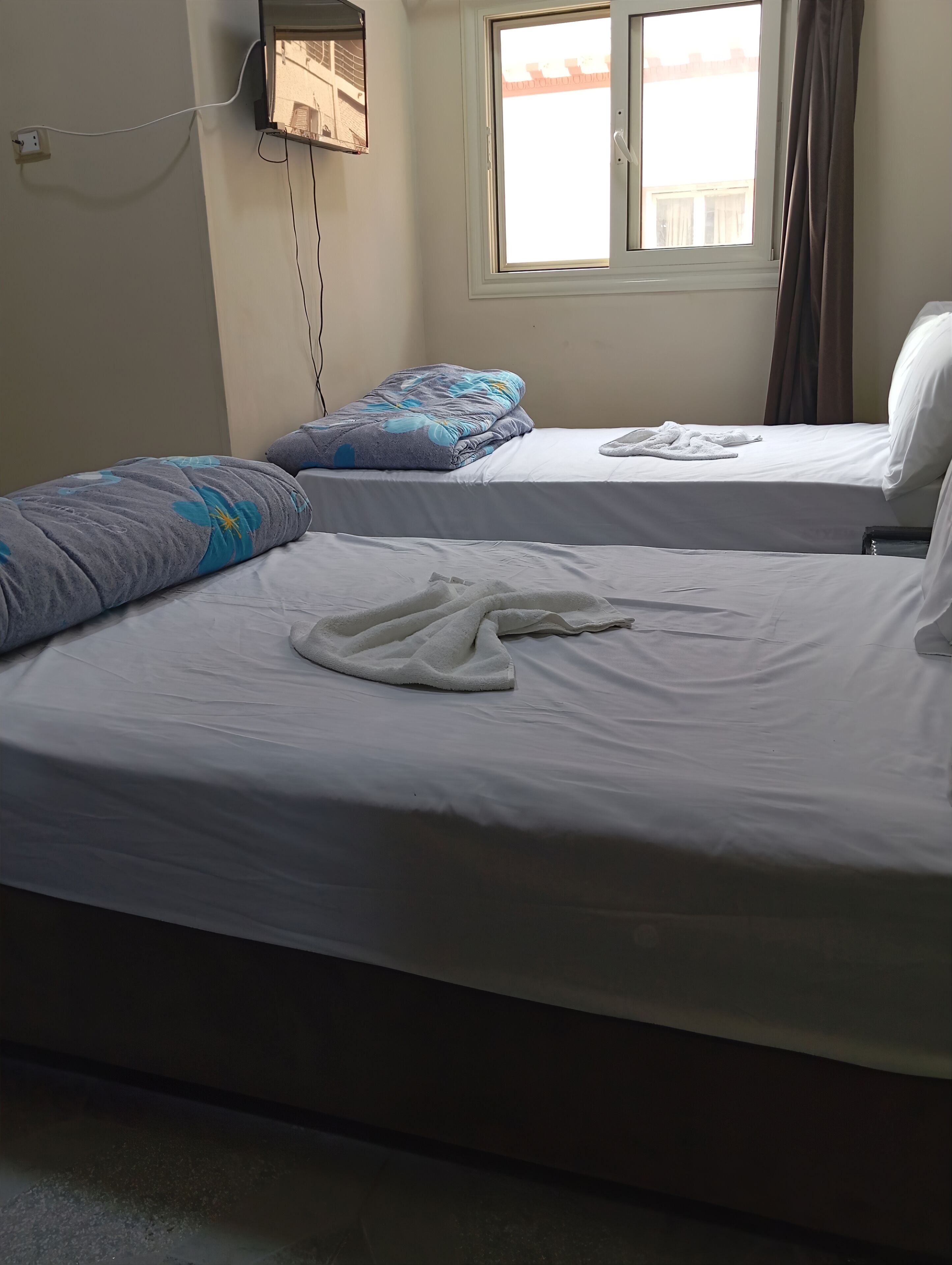 A hotel room with two beds, each with a pillow and a blanket. There is a window with curtains, a wall-mounted TV, and a power outlet.