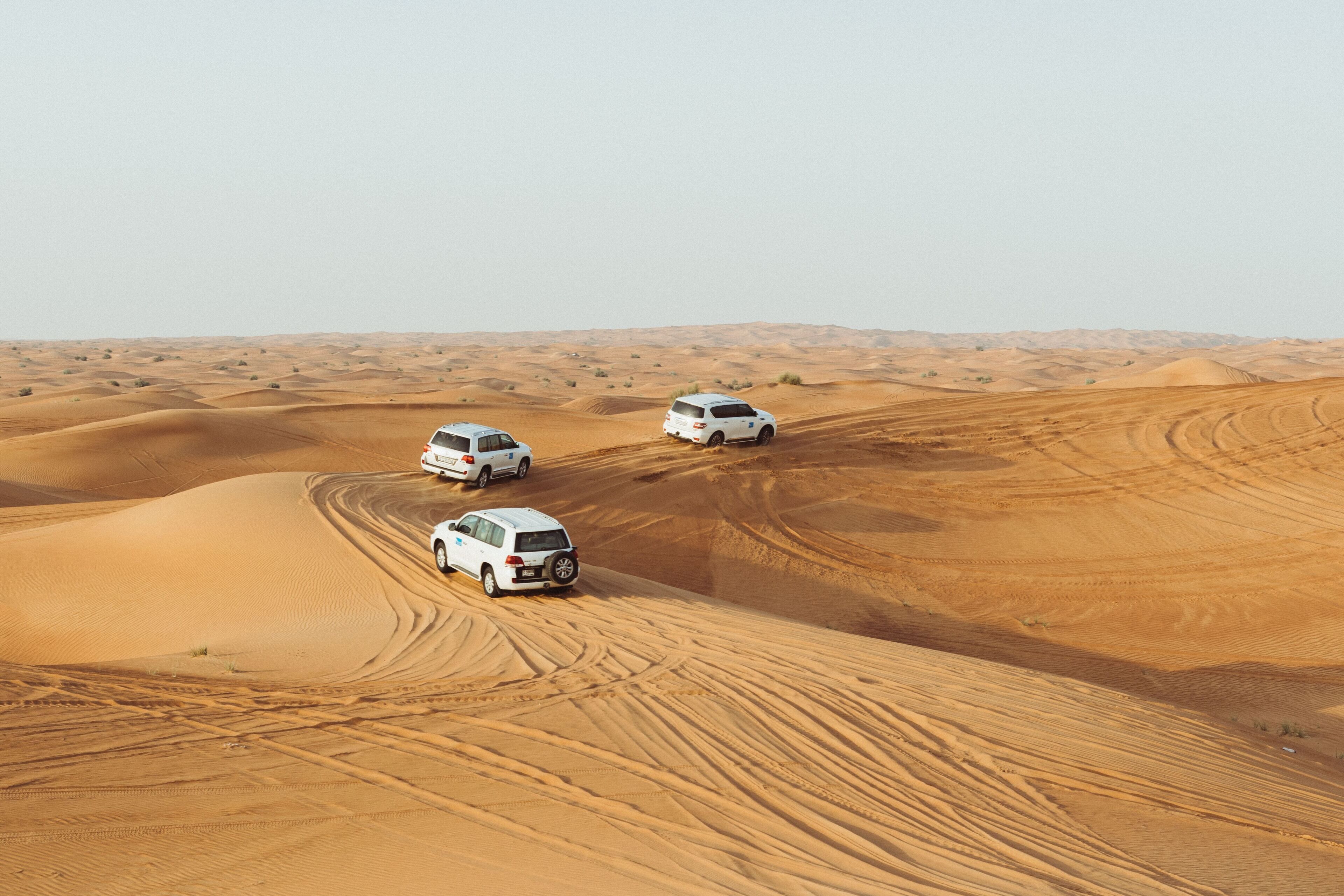Three white SUVs driving on a sandy dune in a desert landscape.