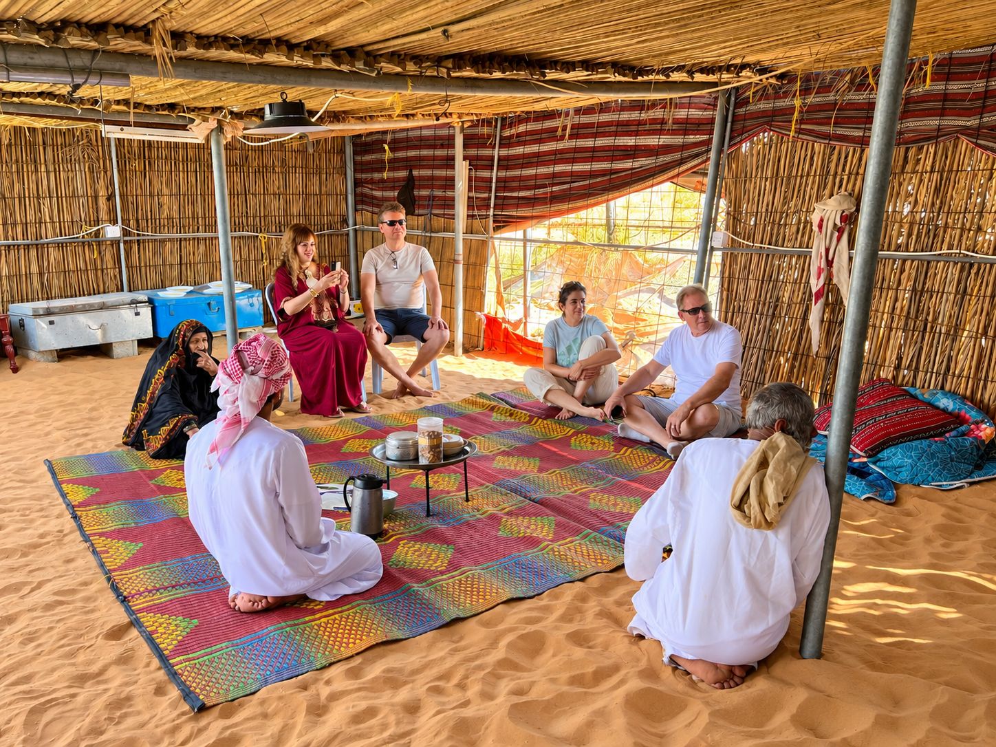 A group of people sitting on colorful mats under a thatched roof, with a metal table in the center.