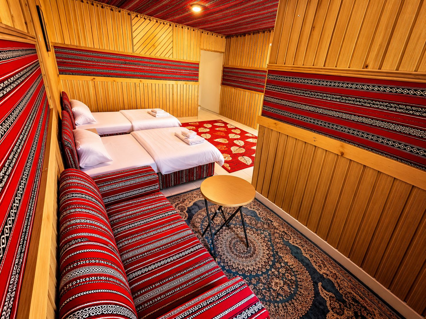A room with wooden paneling, a bed with red and white patterned bedding, a small round table, and a patterned rug.