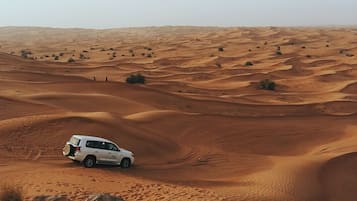 A white SUV driving through a desert landscape with undulating sand dunes.