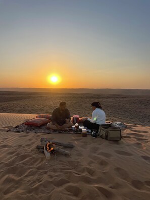 Two people sitting on a blanket in a desert at sunset with a fire and various items around them. - Al Salam Camp (Bidiya)