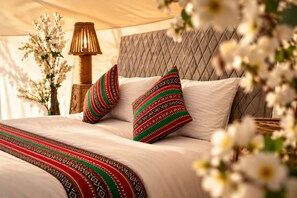 A bed with a patterned bedspread and pillows, a lamp, and a vase with flowers. - Al Salam Camp (Bidiya)