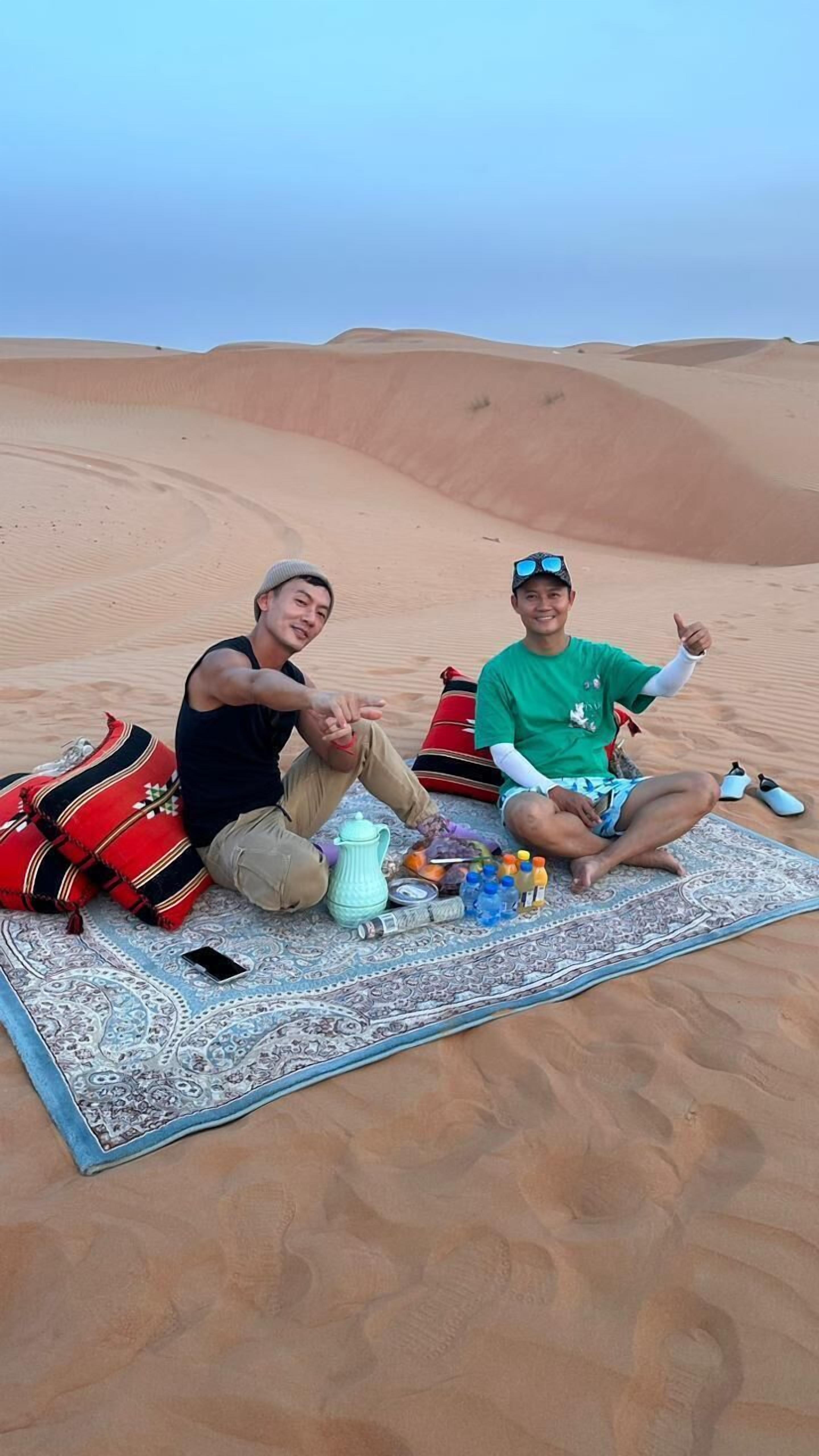 Two individuals sitting on a patterned rug in a desert setting with sand dunes in the background.