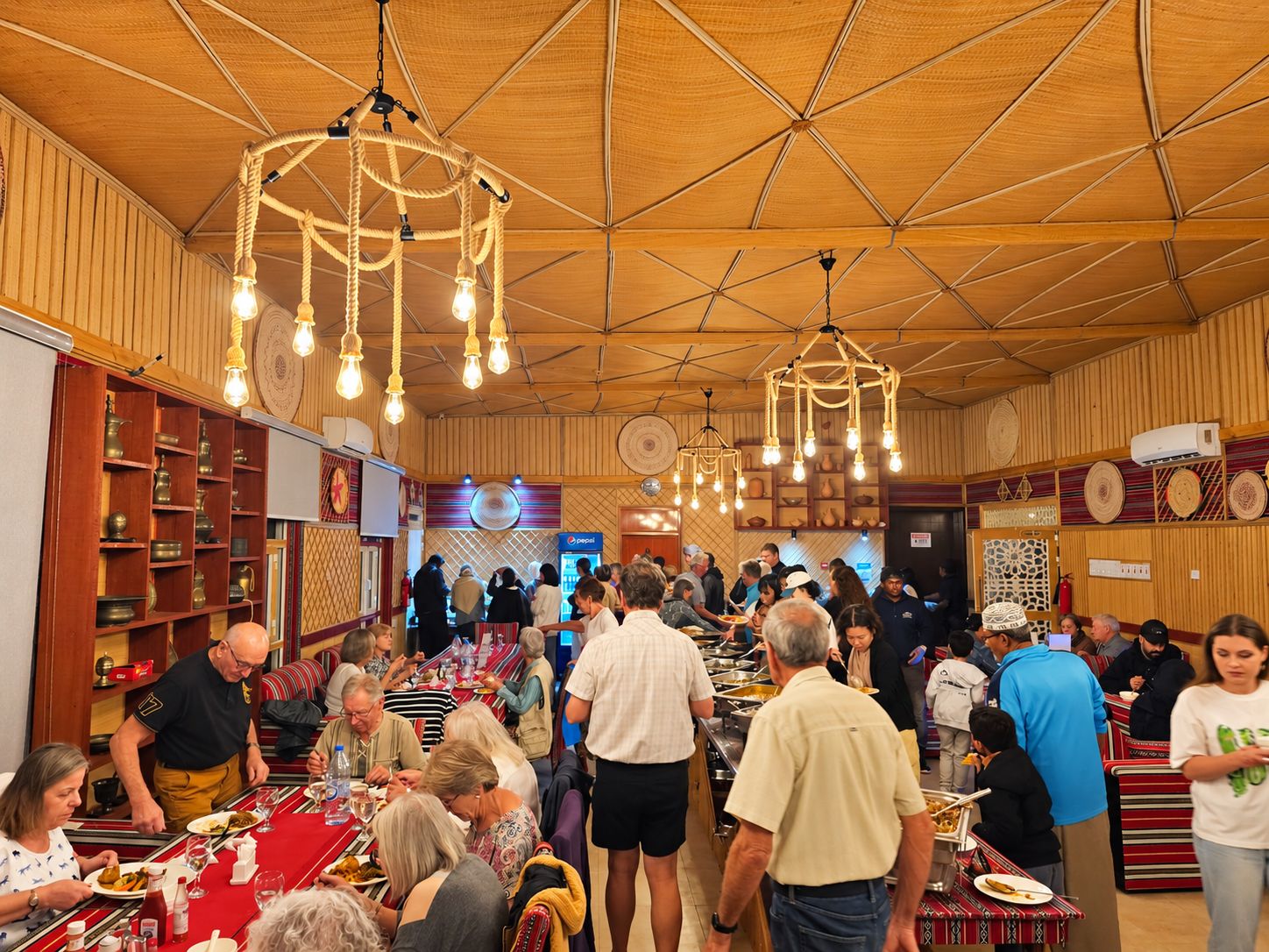 A crowded indoor dining area with wooden walls and ceiling, multiple hanging light fixtures, and a buffet setup.