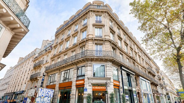Un edificio en esquina de varios pisos, con comercios en la planta baja, incluyendo una pizzería y un restaurante.